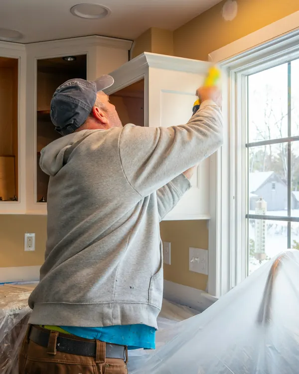 A handyman in a cap and gray sweatshirt sealing the trim above a kitchen window, with the counters covered in plastic