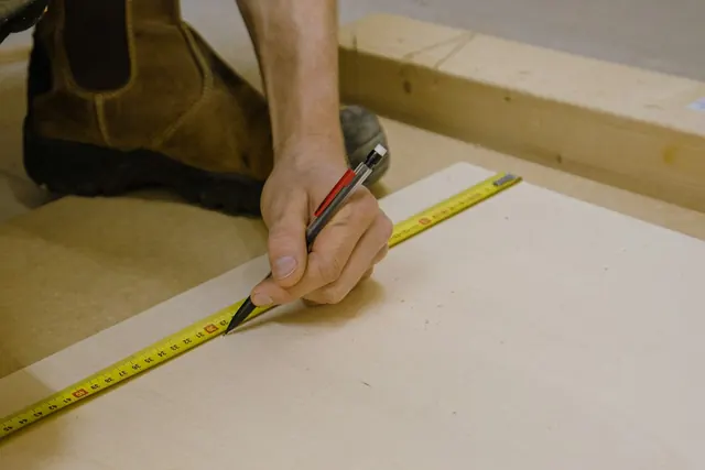 A hand marking a sheet of plywood with a pencil along a tape measure