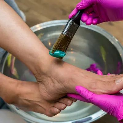 A gloved technician brushing a soft mask onto a client's foot over a steel foot bath